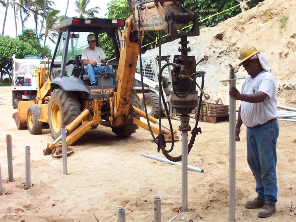 Structural Systems Hawaii crew installing helical piers using backhoe-mounted drive head at beachfront site