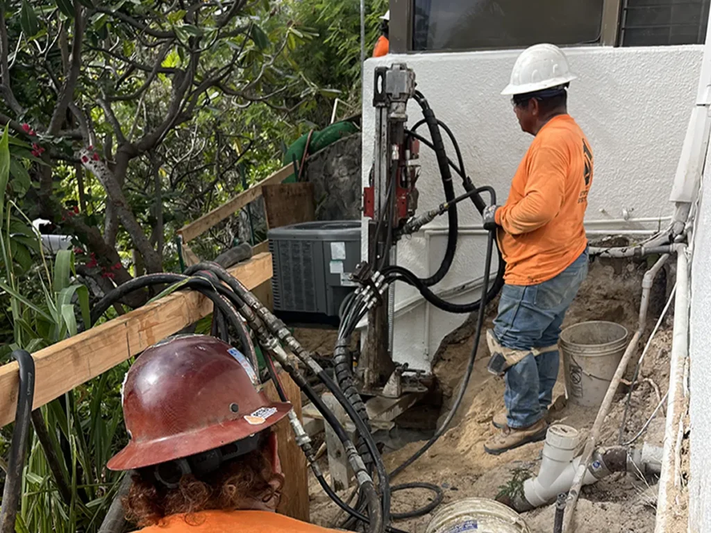 Structural Systems Hawaii technicians operating hydraulic drill rig for foundation repair beside home.