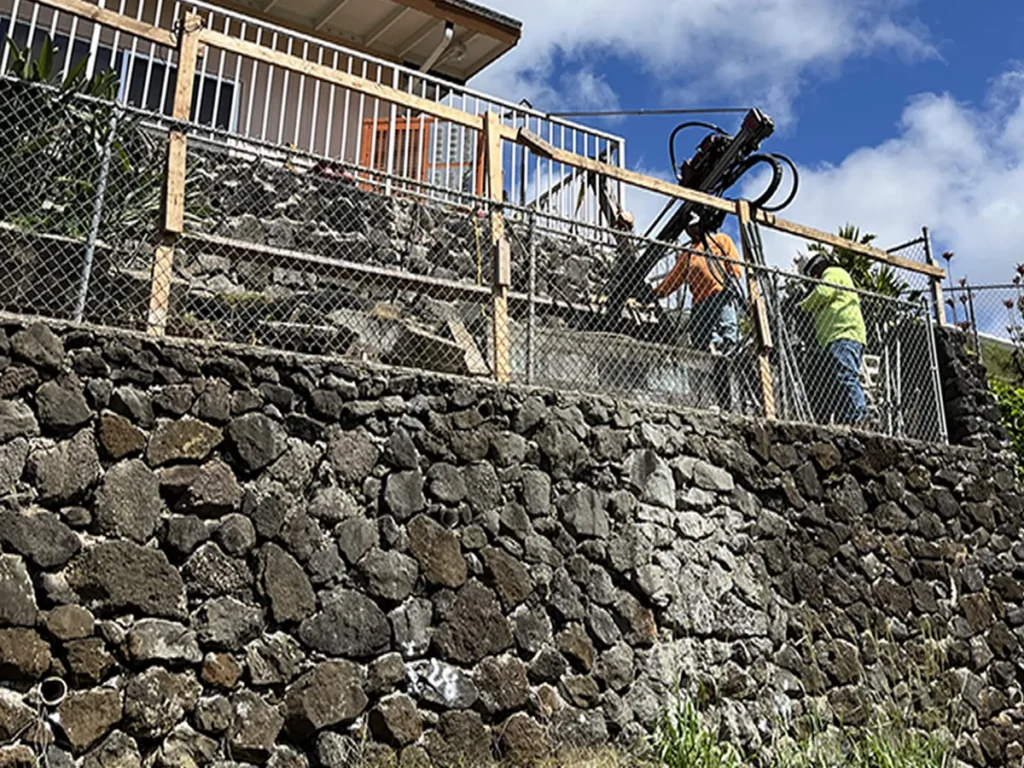 Structural Systems Hawaii technicians drilling stone retaining wall for repair above residential home